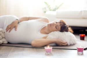 Healthy Pregnancy Yoga and Fitness. Young pregnant yoga woman resting after working out in living room interior. Pregnant model lying in prenatal Shavasana (Corpse, Dead Body Pose). Focus on candle